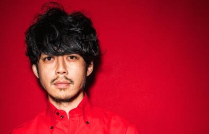 赤い背景を背にして立つ、黒髪でパーマヘアの男性