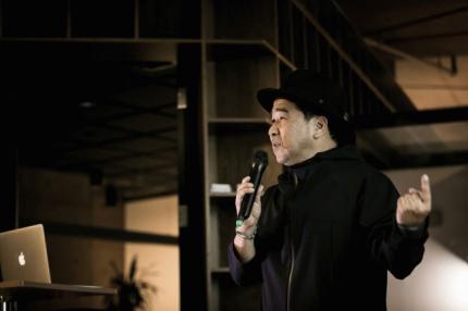 黒いハットと服を着用しマイクを持って話す男性